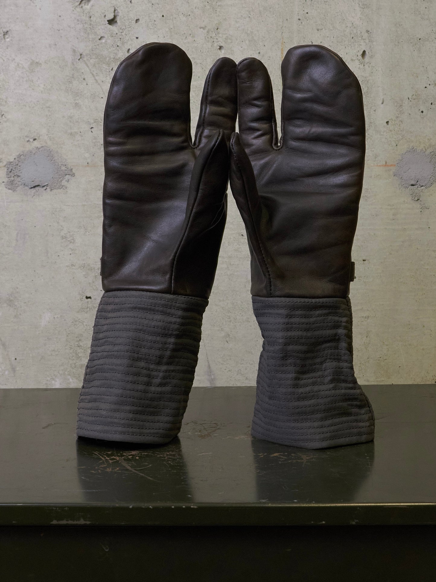 A pair of dark brown leather tabi gloves with ribbed gray fabric cuffs, standing upright on a dark surface against a textured concrete wall.
