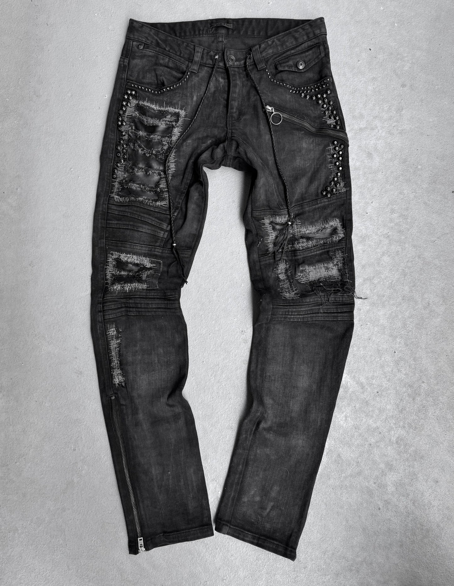KMRii Early 00s Goatskin Patches Studded Crush Flare-zip Denim in black, US 29-30 waist. Features multiple rips, patches, studs, zippers, and metal ring details with slight fading for an edgy look. Laid flat on a light surface.