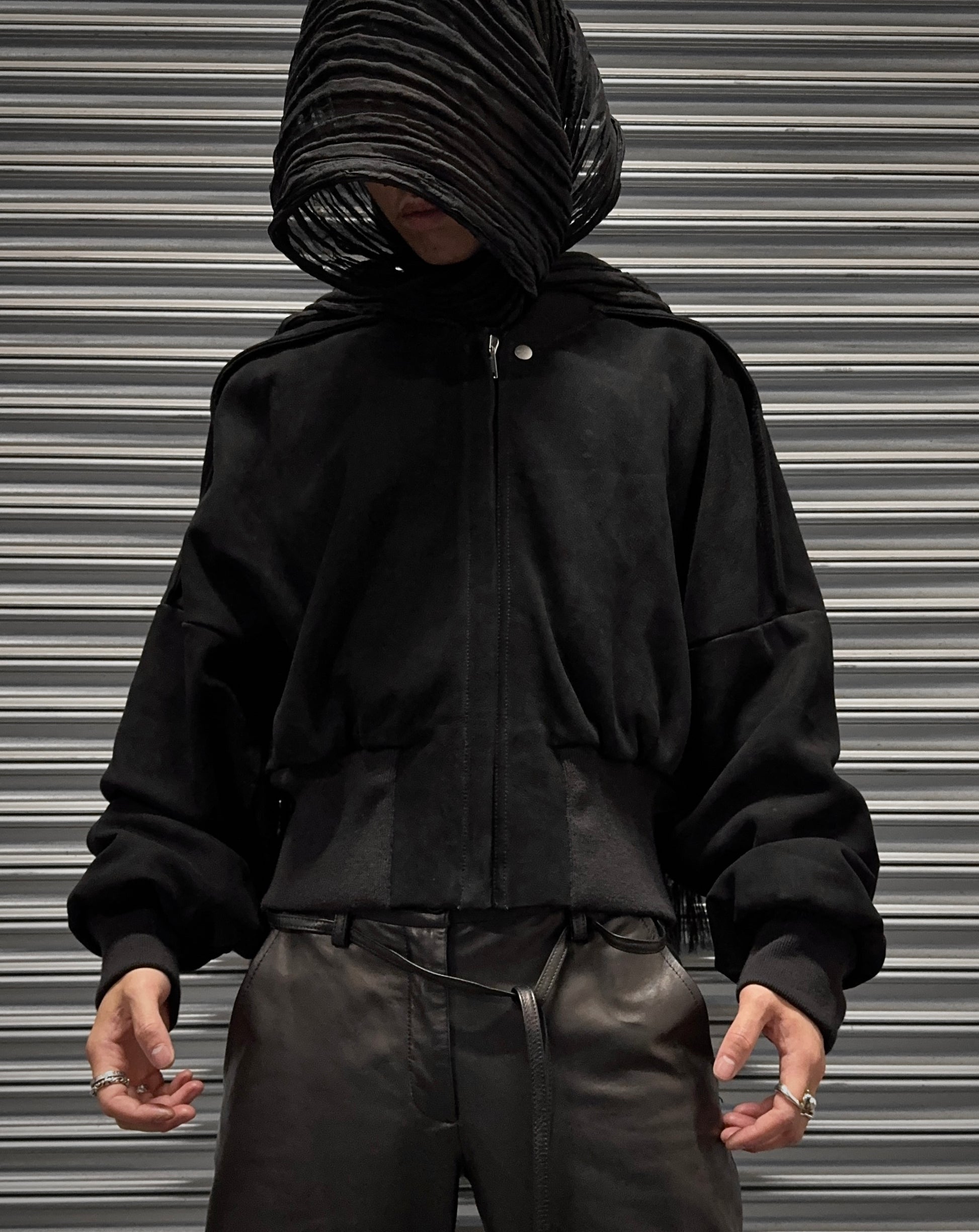 A person stands before a metal shutter in the Rick Owens SS24 “LIDO” Nubuck Leather Cropped Flight Bomber Jacket, loose black pants, and a textured black head covering, their ring-adorned hands finishing the avant-garde look.
