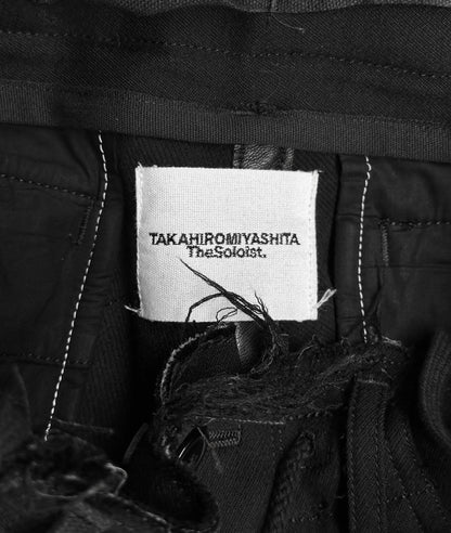 Close-up of a clothing label on black pants, featuring TakahiroMiyashita TheSoloist. in bold text against white. These avant-garde AW15 Calf-trimmed Wool Bondage Straps Cargo Pants have black stitching and artfully frayed edges near the label.