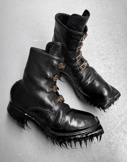 A pair of Carol Christian Poell ‘AM/2759SP-IN CORSS-PTC’ Horse Leather Buckle Drip Boots with distinct metal fastenings and claw-like spiked soles, showcased on a light gray surface.