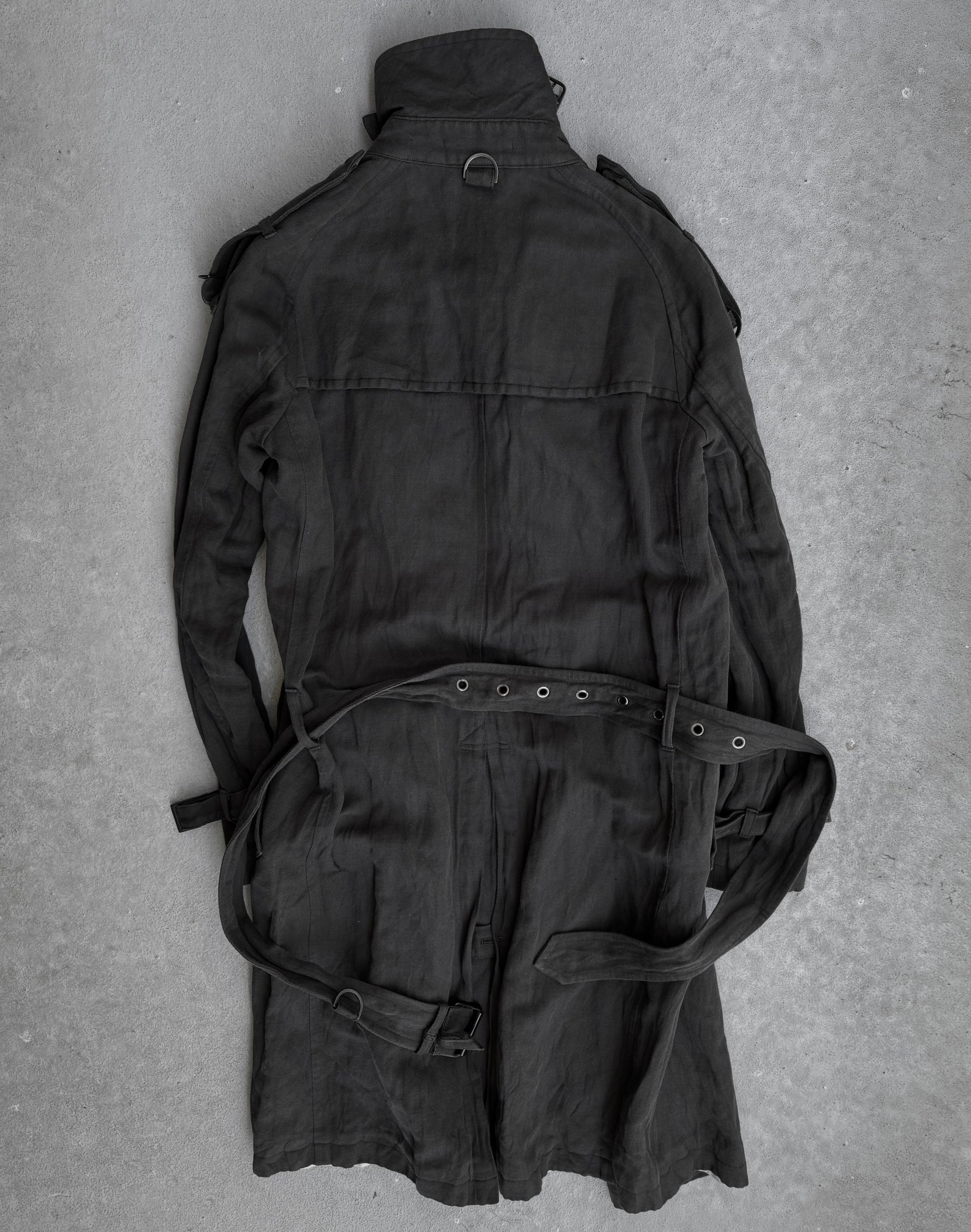 The ATTACHMENT by Kazuyuki Kumagai Raglan Doctor Trench Coat is laid flat on a gray concrete surface, displaying its back and sleeves—a sleek piece from Attachment by Kazuyuki Kumagai.