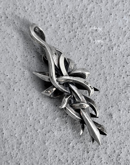 Close-up of M’s COLLECTION (BLESS) Early 00s Silver Arabesque Cross Pendant by BLESS, featuring intertwining vine-like designs on a textured gray background. Its dynamic and elegant curved, pointed elements reflect the spirit of sustainability.