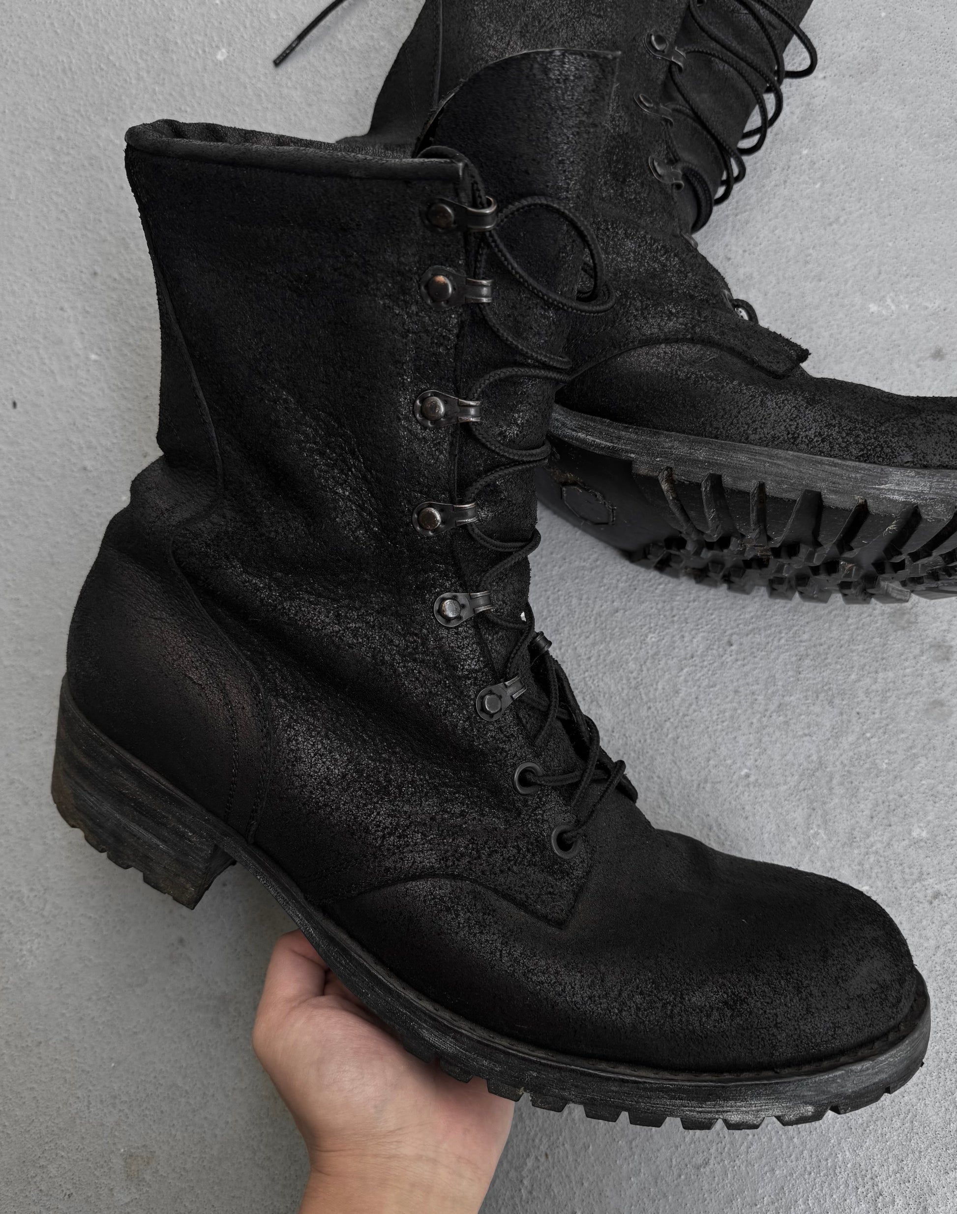 A hand holds an ATTACHMENT by Kazuyuki Kumagai Reversed Cow Leather Lace-up Boot with a rugged sole, while another similar boot from Attachment by Kazuyuki Kumagai rests on a gray floor in the background.