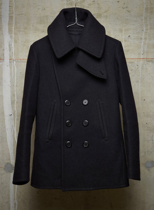 A black double-breasted peacoat with a wide collar, hanging on a wire hanger against a textured concrete wall.