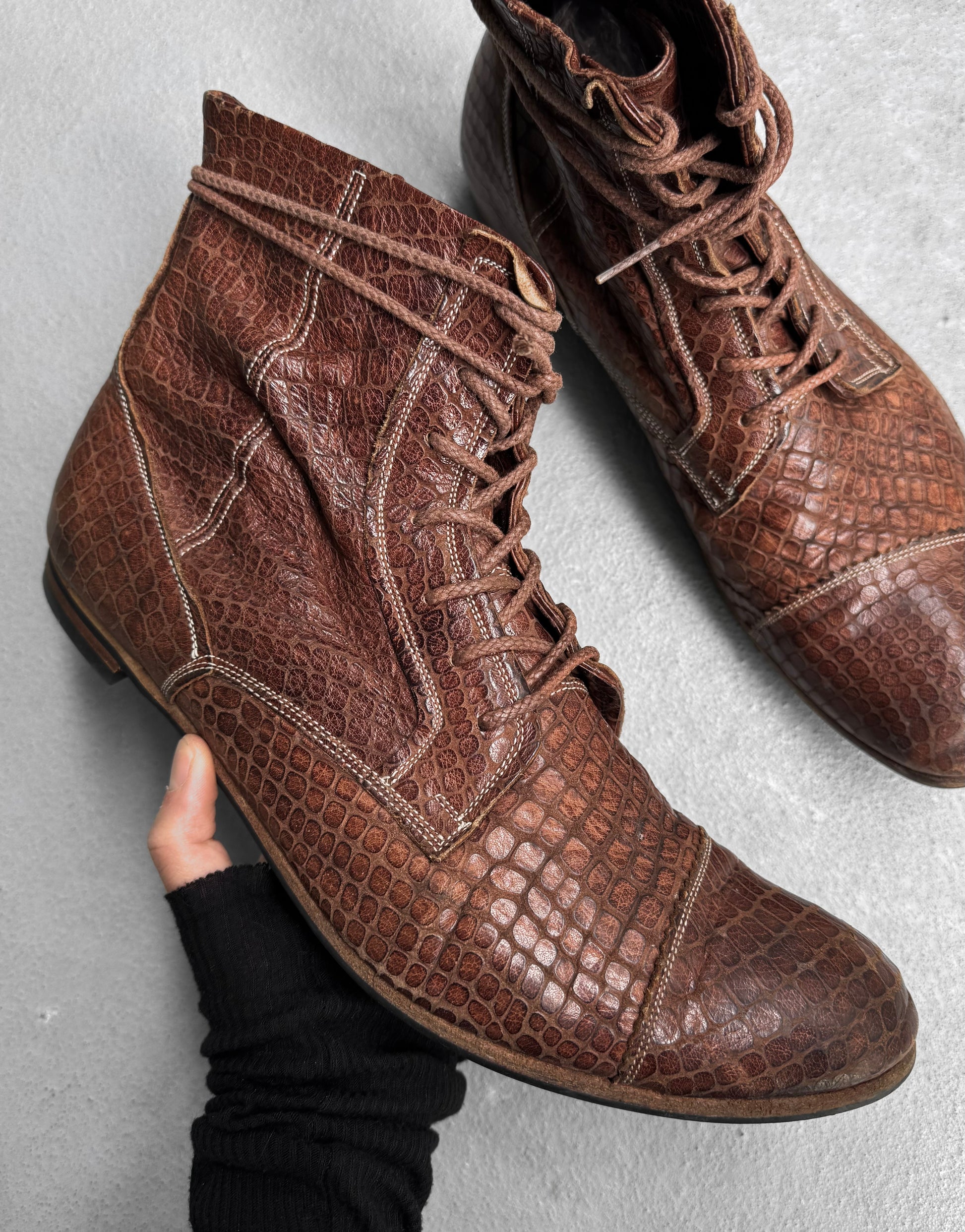 A hand holds the Mihara Yasuhiro Early 00s Python Embossed Cow Leather Pointy Toe Boot, while its matching pair rests beside it on a light gray surface.