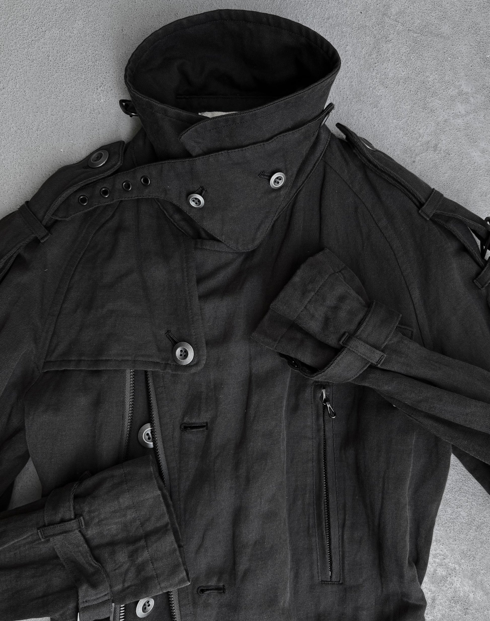A dark grey ATTACHMENT by Kazuyuki Kumagai Raglan Doctor Trench Coat with a high collar, multiple buttons, epaulets, strap details, and a zippered pocket lies flat on a light grey surface—a refined menswear piece.