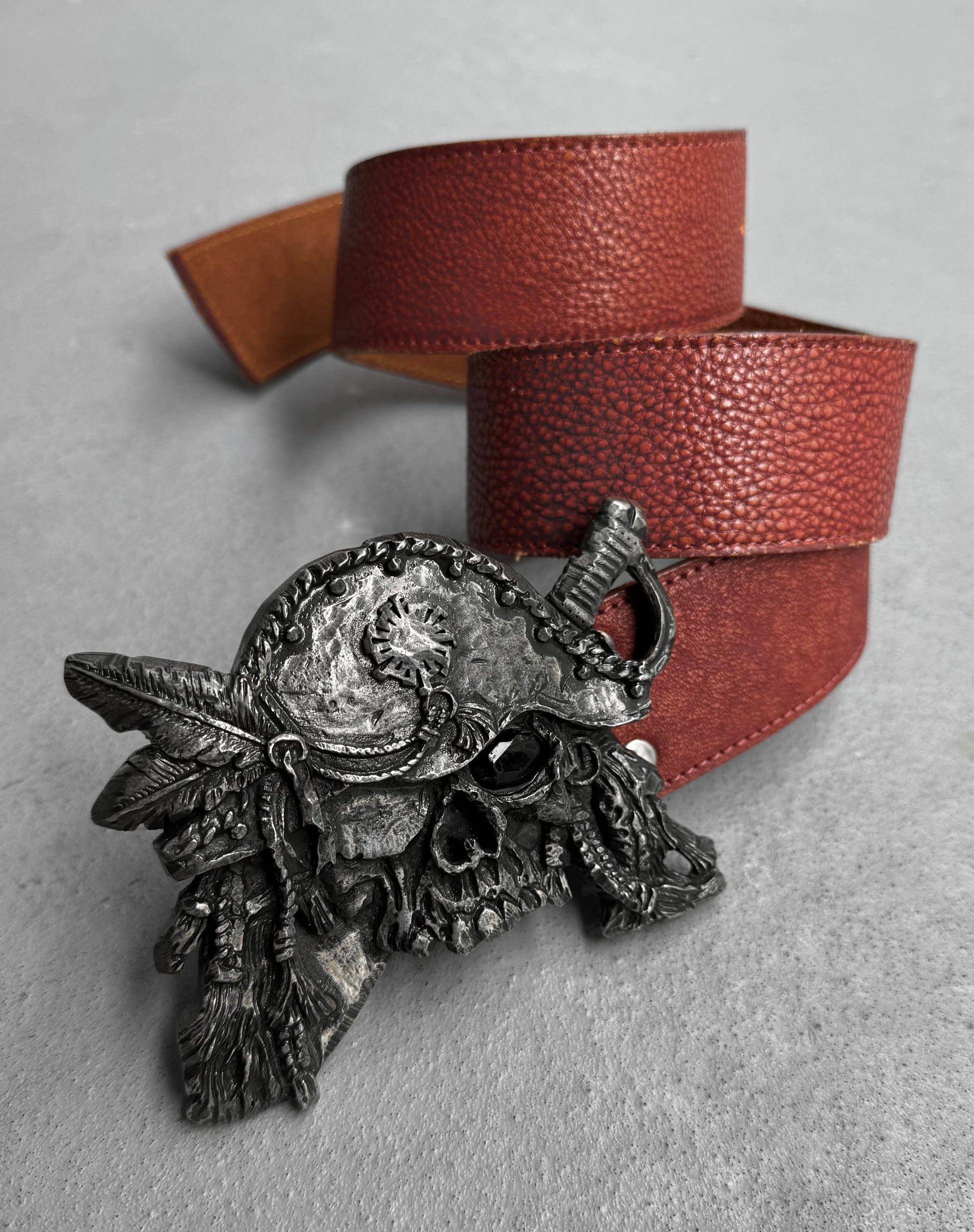 The Tornado Mart Early 00s Pirate Silver Buckle Leather Belt, inspired by Japanese men’s fashion, features a brown leather strap and a detailed pirate skull buckle with a tricorn hat, feathers, and eye patch, shown on a light gray surface.