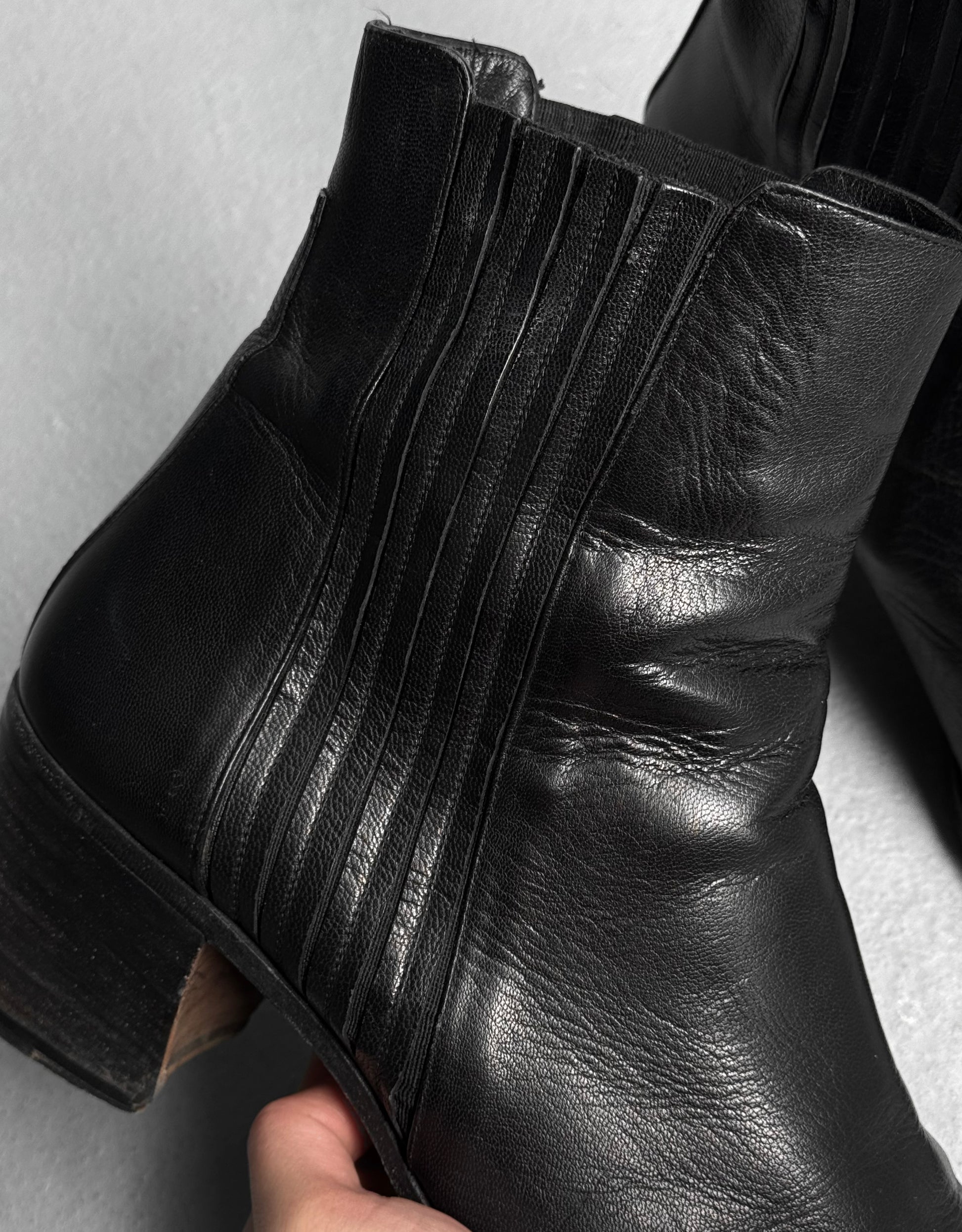 A hand holds the Saint Laurent Paris YSL Rive Gauche SS04 by Tom Ford ‘Johnny’ black leather ankle boot, featuring vertical stitched detailing and a 40mm low stacked heel, against a light neutral background.
