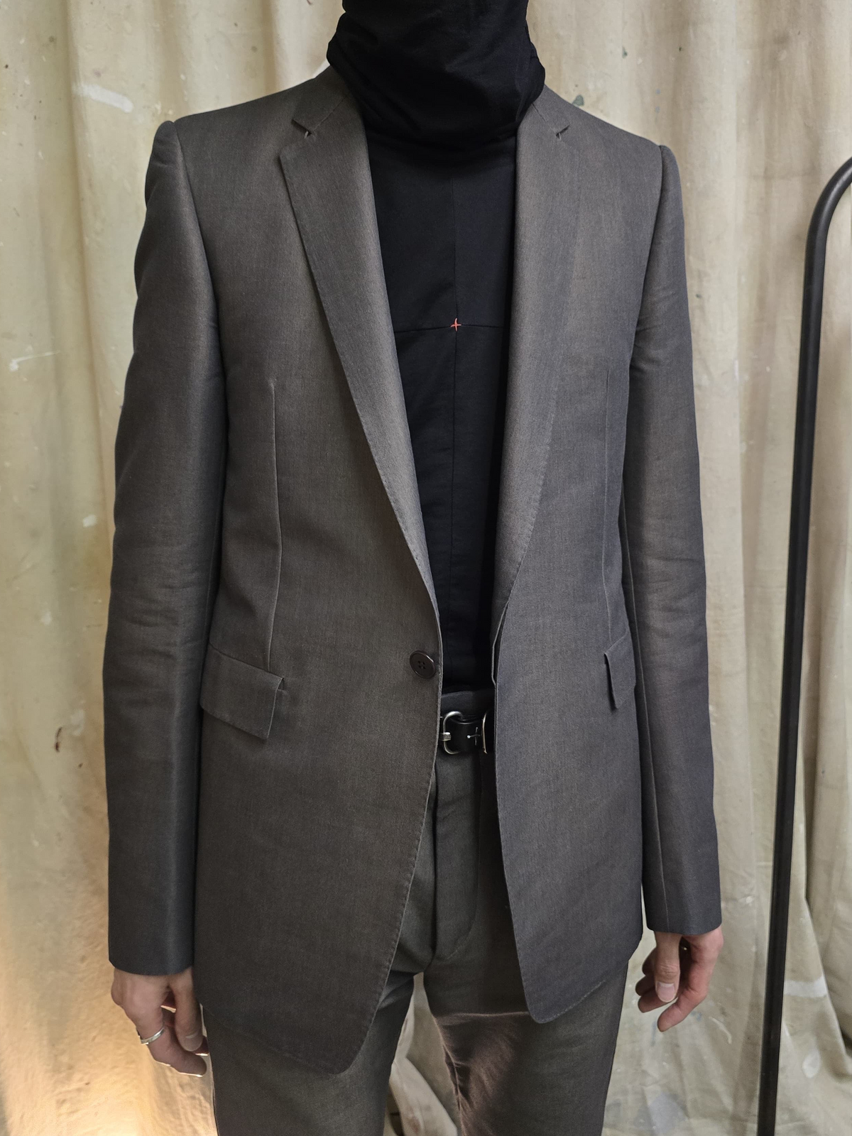 A man wearing a tailored gray blazer with matching gray trousers and a black turtleneck, standing against a beige fabric backdrop.