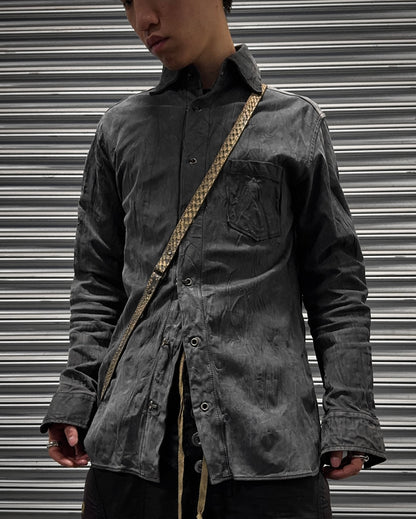 A person in a faded black Kyoji Maruyama Early 00s Washable Wrinkle Nubuck Leather Shirt with a front pocket and tan crossbody strap stands before a metallic, striped backdrop, their face partially visible, channeling Japanese minimalism.
