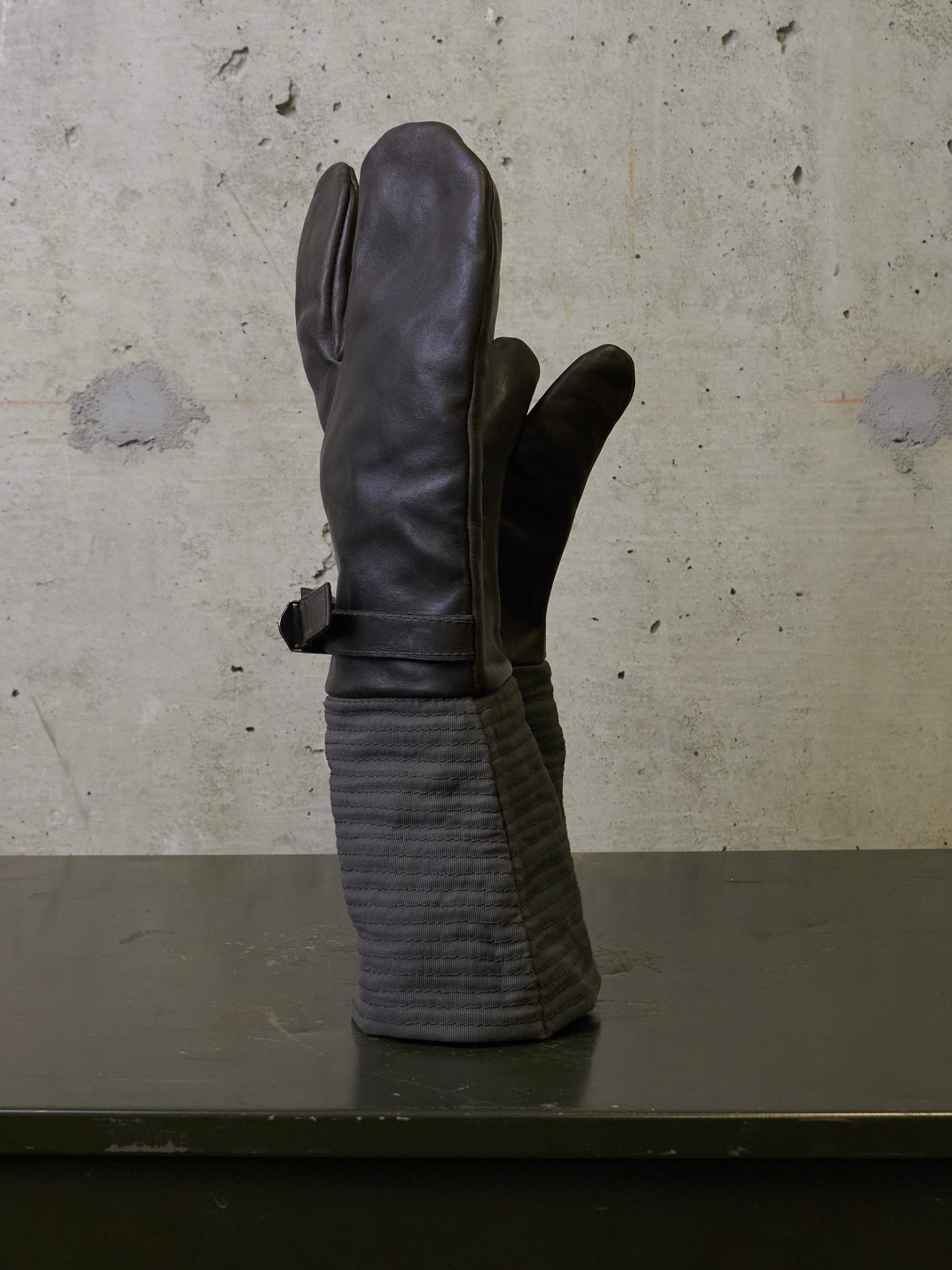 A pair of dark brown leather mittens with ribbed fabric cuffs, standing upright on a dark surface against a textured concrete wall.