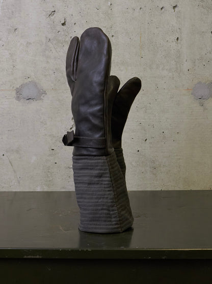 A pair of dark brown leather mittens with ribbed fabric cuffs, standing upright on a dark surface against a textured concrete wall.