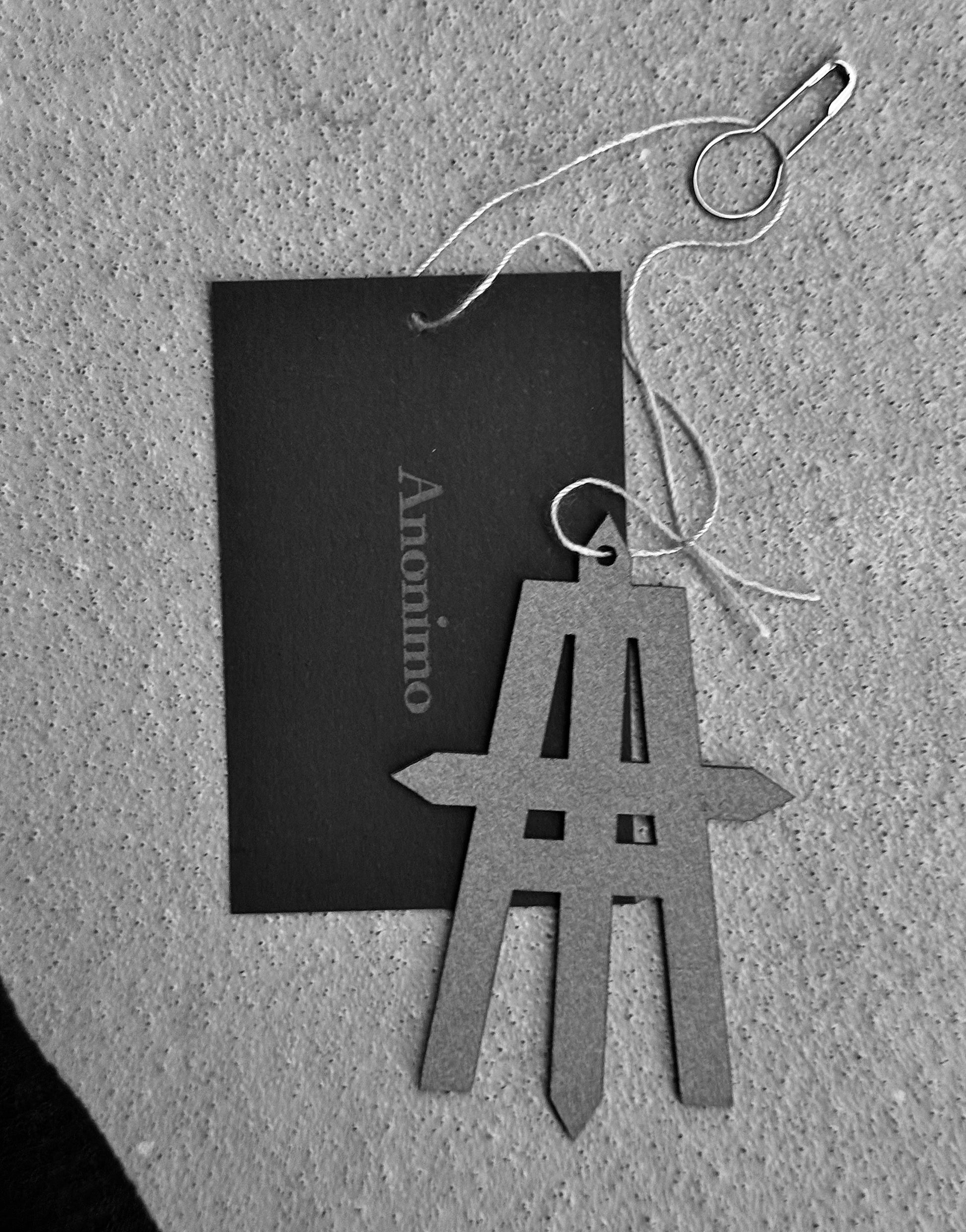 A Maison Anonimo tag rests on a textured surface with a gray geometric pendant and string, beside a safety pin. This black-and-white image features the MAISON ANONIMO Cross-Dart Cotton-bamboo Hat—ideal for stylish accessories.
