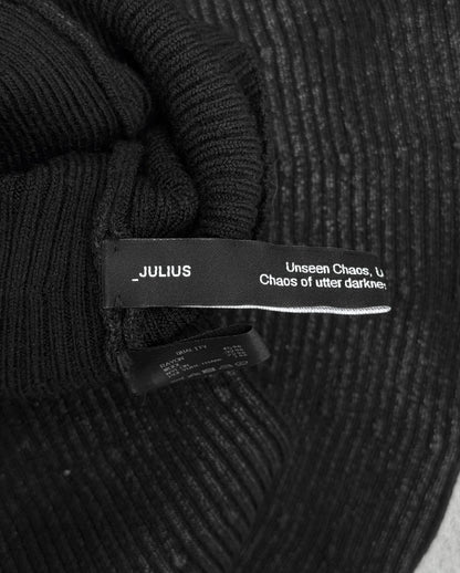 A close-up of DRIEW’s Julius_7 AW24-25 “rift;” Curved Wax Coated Gloves in flawless black ribbed fabric, featuring a “_JULIUS” label with partially visible text: “Unseen Chaos… utter darknes…”. One size fits most.