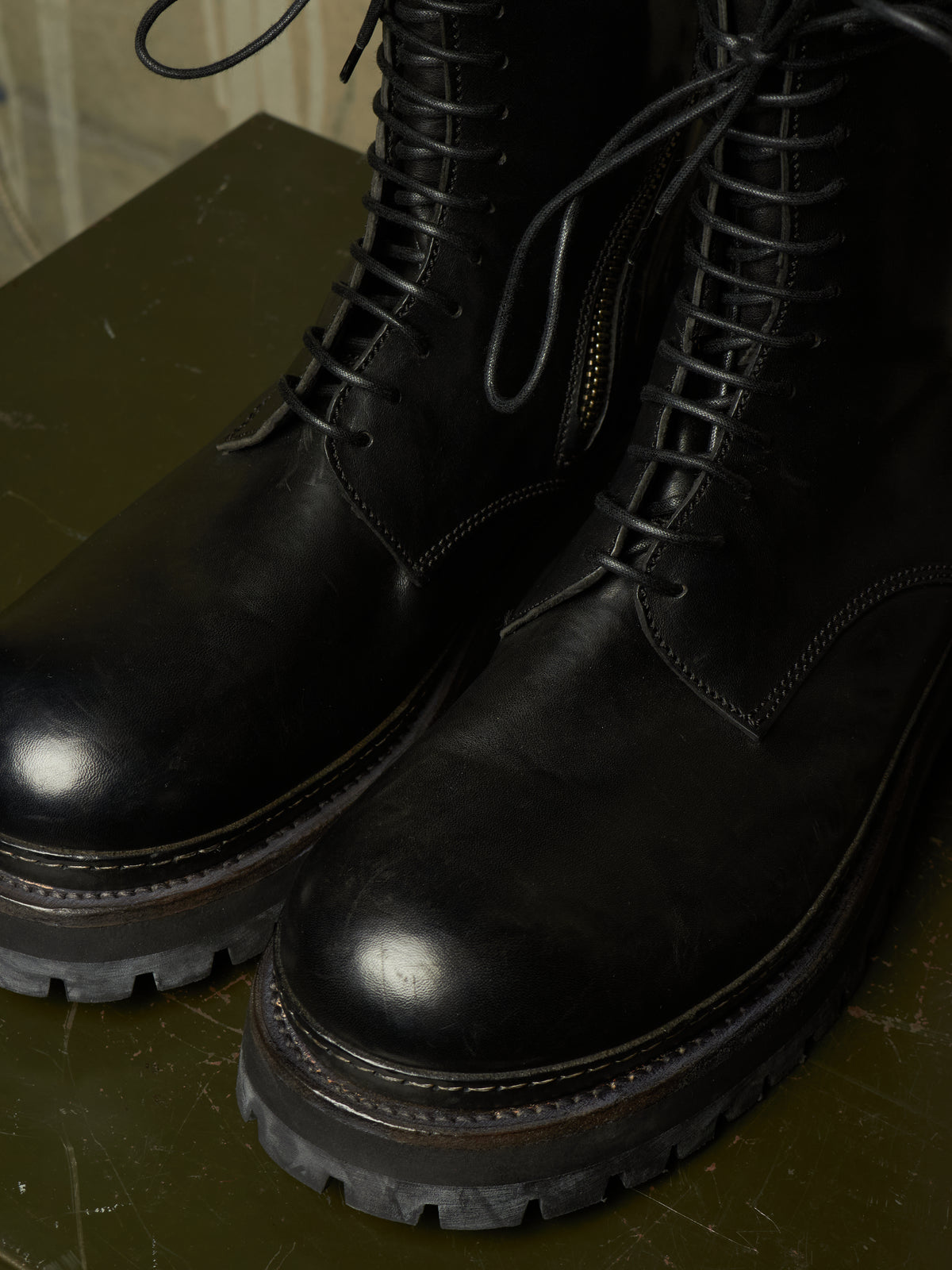 A close-up of a pair of black leather lace-up boots with rugged lug soles, resting on a dark olive green surface.