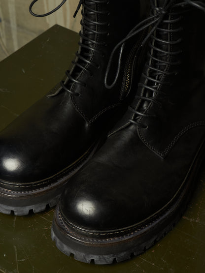 A close-up of a pair of black leather lace-up boots with rugged lug soles, resting on a dark olive green surface.