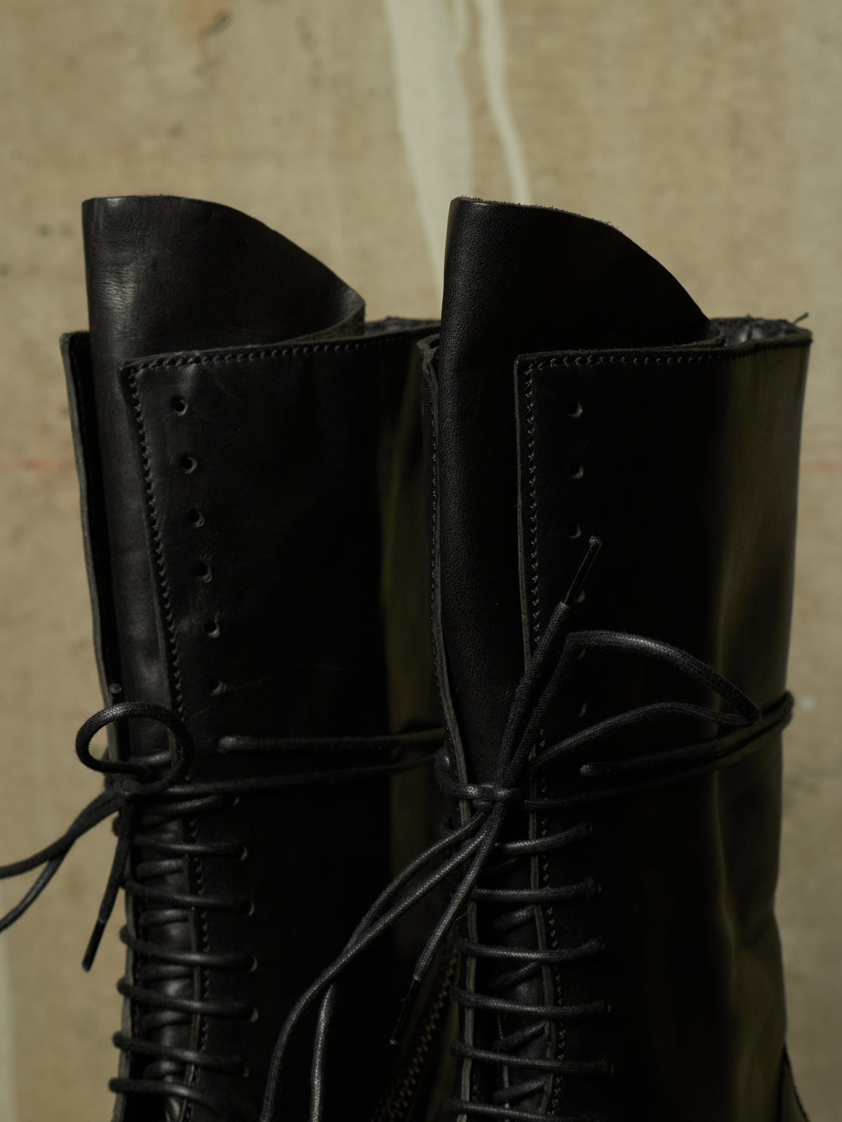 A close-up of a pair of black leather lace-up boots with pointed toes, set against a textured beige background.