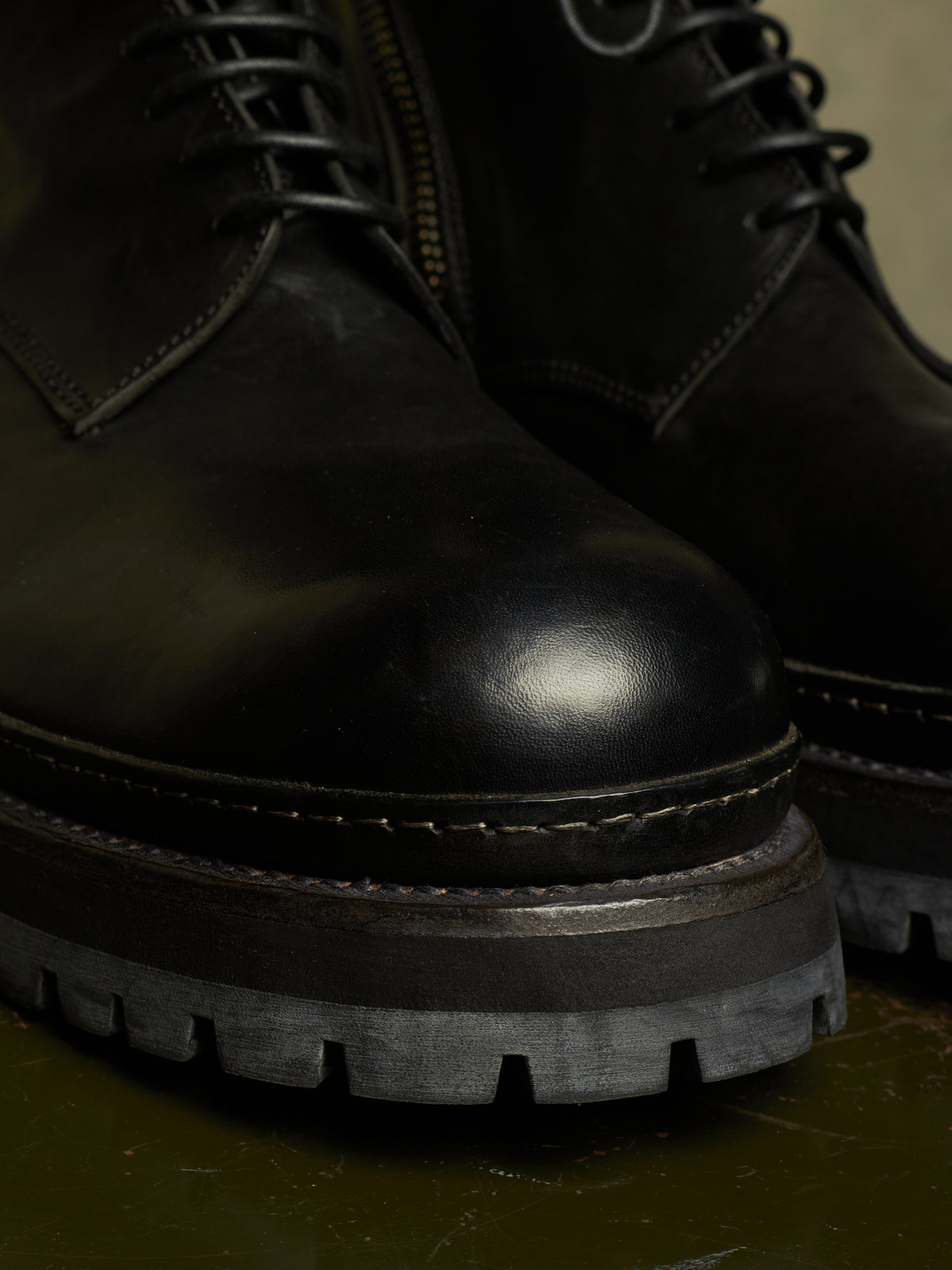 Close-up of a black leather lace-up boot with a rugged sole, showing the toe, laces, stitching, and part of the second boot behind it.