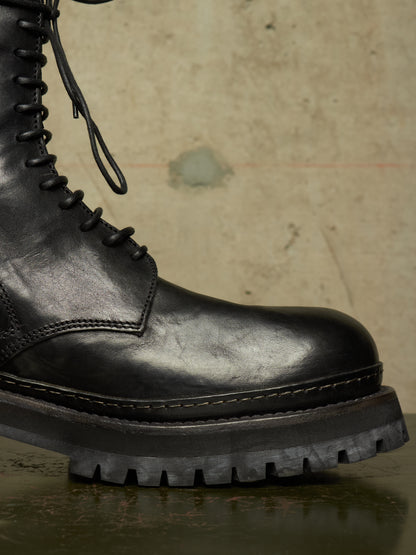 Close-up of a black leather lace-up boot with a chunky lug sole, shown against a textured concrete background.