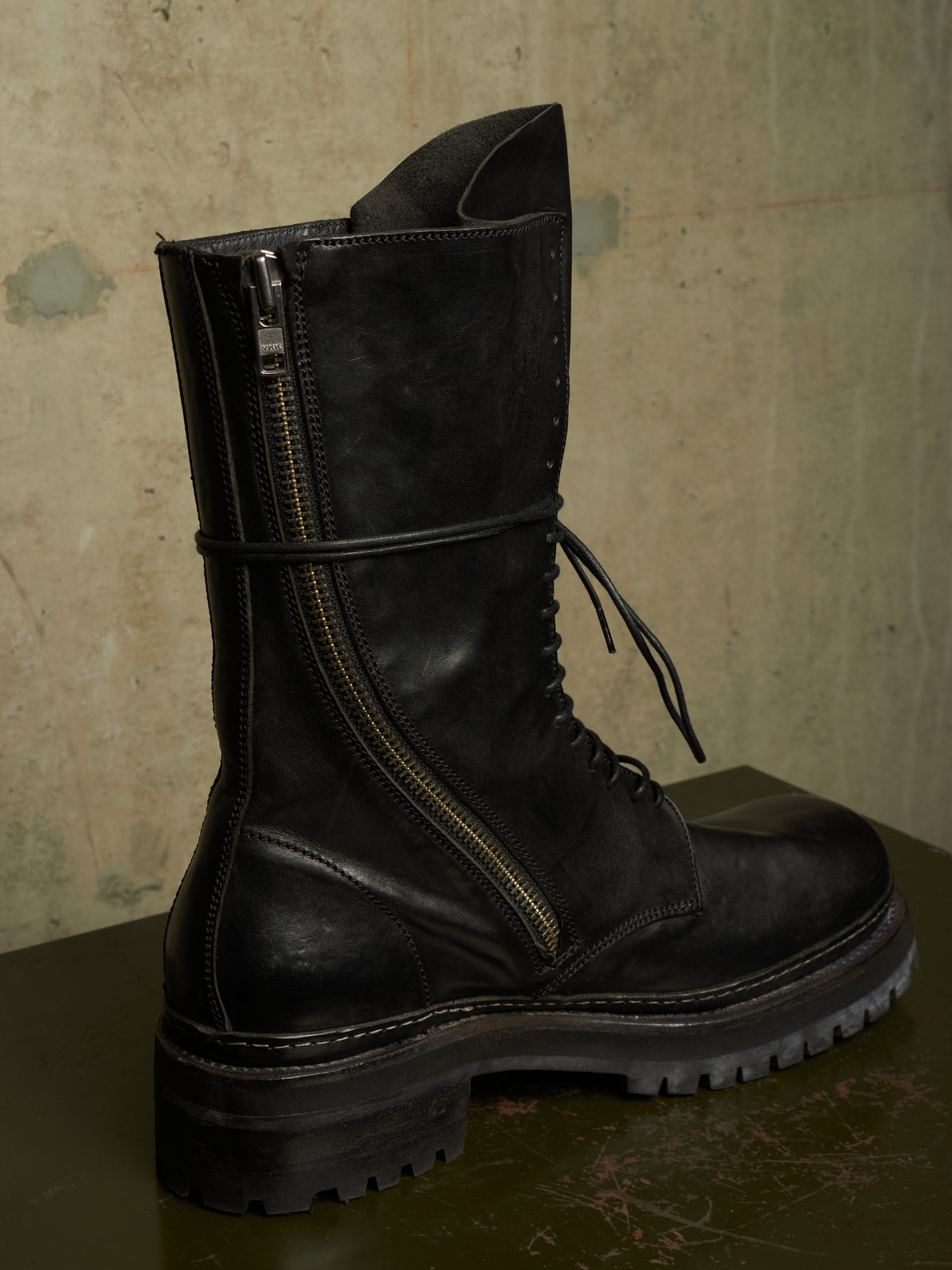 A black leather lace-up combat boot with a prominent side zipper and a thin black strap around the ankle, resting on a dark surface against a textured concrete wall.