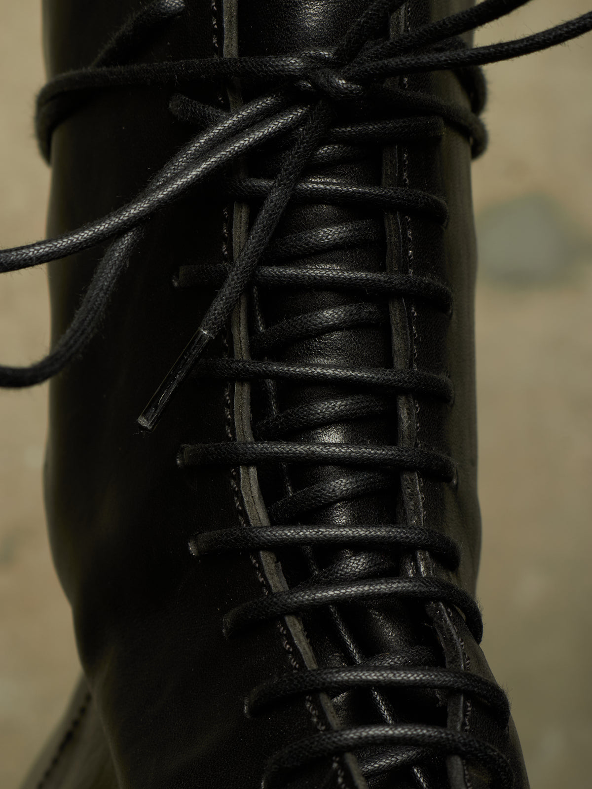 Close-up of a black leather lace-up boot with black laces threaded through metal eyelets, set against a blurred beige background.