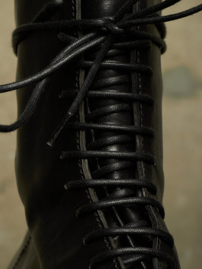 Close-up of a black leather lace-up boot with black laces threaded through metal eyelets, set against a blurred beige background.