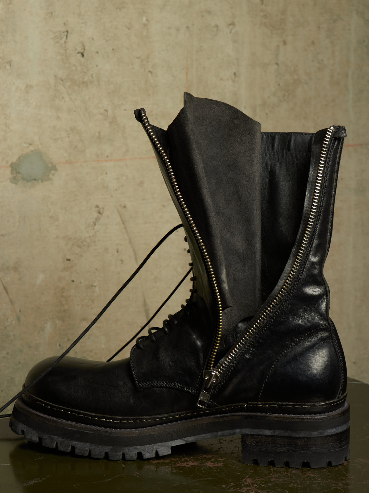 A black leather lace-up combat boot with a prominent side zipper and a black fabric sleeve extending from the zipper, set against a textured concrete background.