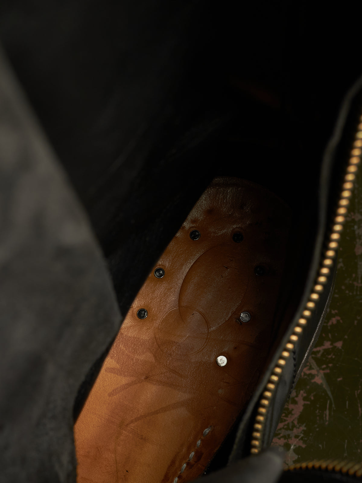 Close-up of a brown leather insole inside a dark bag, showing a gold zipper and small metal rivets.