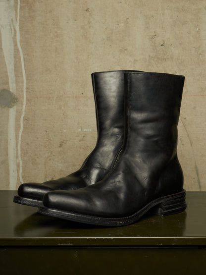A pair of black leather mid-calf boots with a stacked lug sole, displayed on a dark green surface against a textured beige concrete wall.