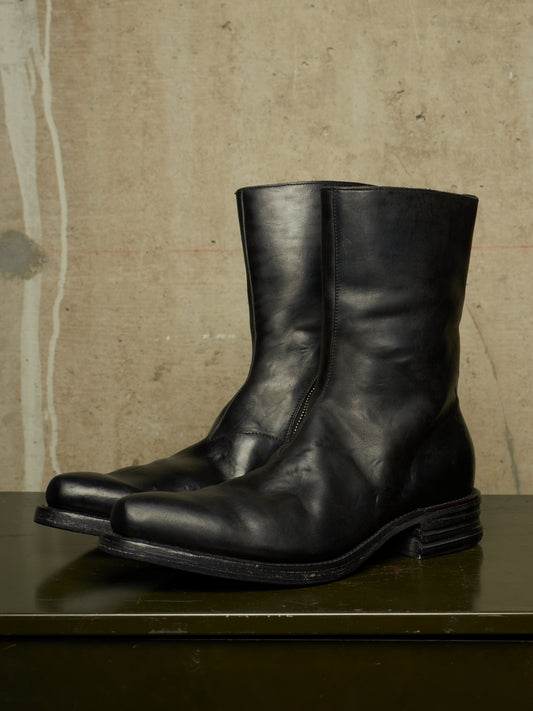 A pair of black leather mid-calf boots with a stacked lug sole, displayed on a dark green surface against a textured beige concrete wall.