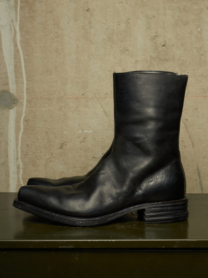 A pair of black leather mid-calf boots with a stacked lug sole, displayed on a dark green surface against a textured beige concrete wall.