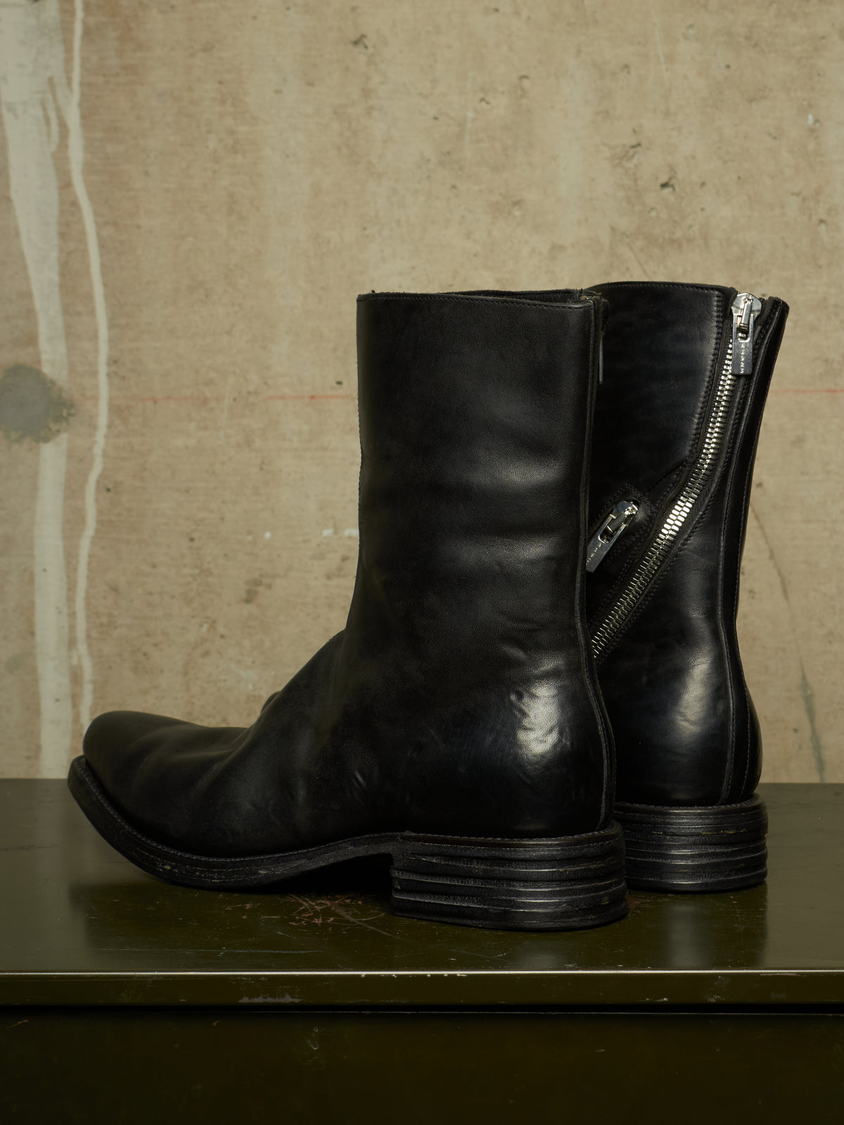A pair of black leather ankle boots with side zippers, displayed on a dark green surface against a textured beige wall.