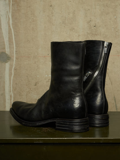 A pair of black leather ankle boots with side zippers, displayed on a dark green surface against a textured beige wall.