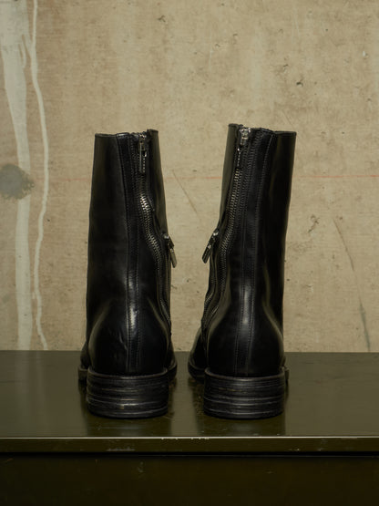A pair of black leather ankle boots shown from the back, featuring side zippers and stacked lug soles, set against a textured beige wall and dark green surface.