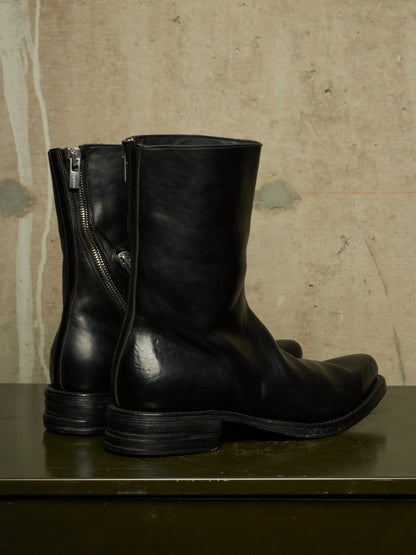 A pair of black leather mid-calf boots with side zippers, displayed on a dark green wooden surface against a textured beige concrete wall.