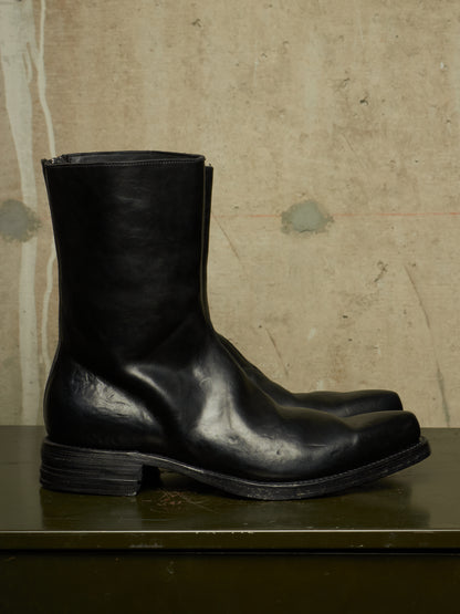 A pair of black leather mid-calf boots with a low stacked heel, displayed on a dark green surface against a textured beige concrete wall.