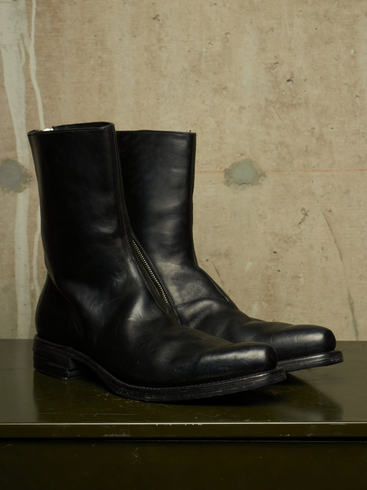 A pair of black leather mid-calf boots with side zippers, displayed on a dark green surface against a textured beige concrete wall.