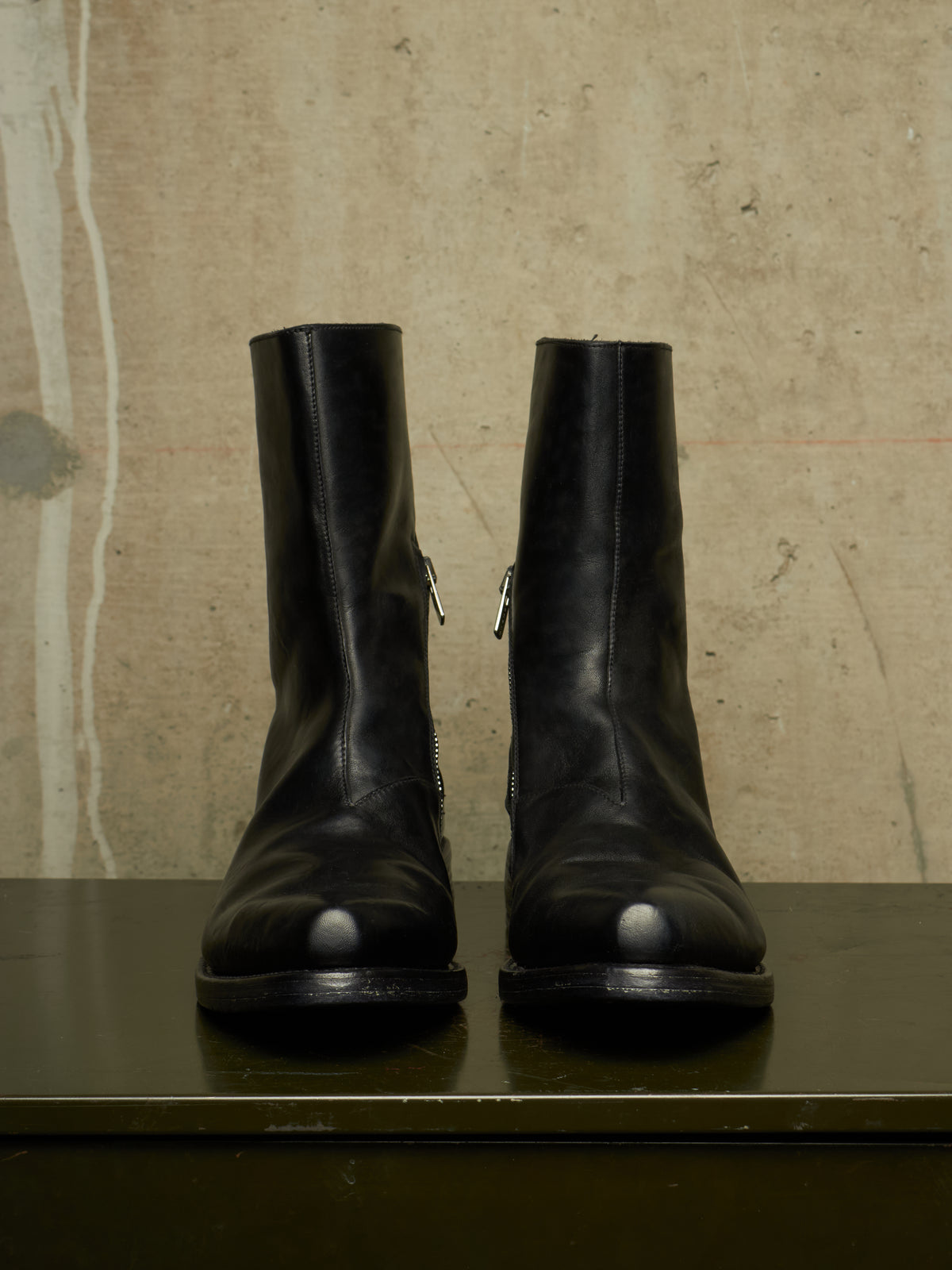 A pair of black leather ankle boots with side zippers, displayed on a dark surface against a textured beige wall.