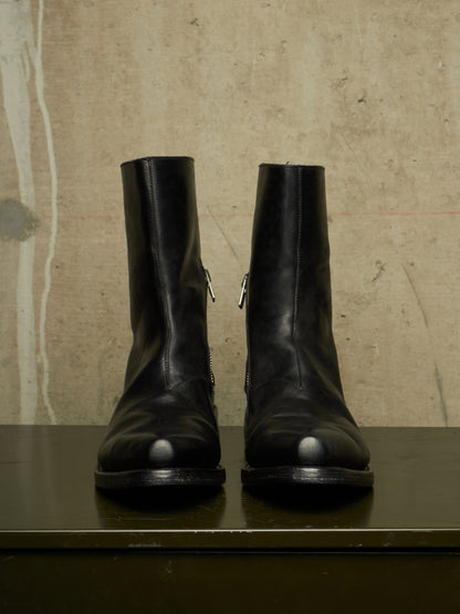 A pair of black leather ankle boots with side zippers, displayed on a dark surface against a textured beige wall.