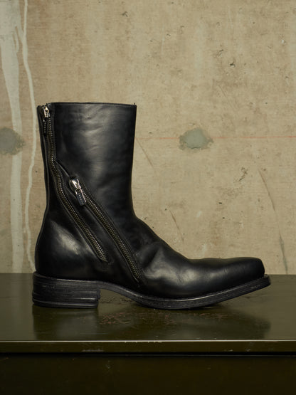 A black leather ankle boot with a side zipper, displayed on a dark green surface against a textured beige wall.