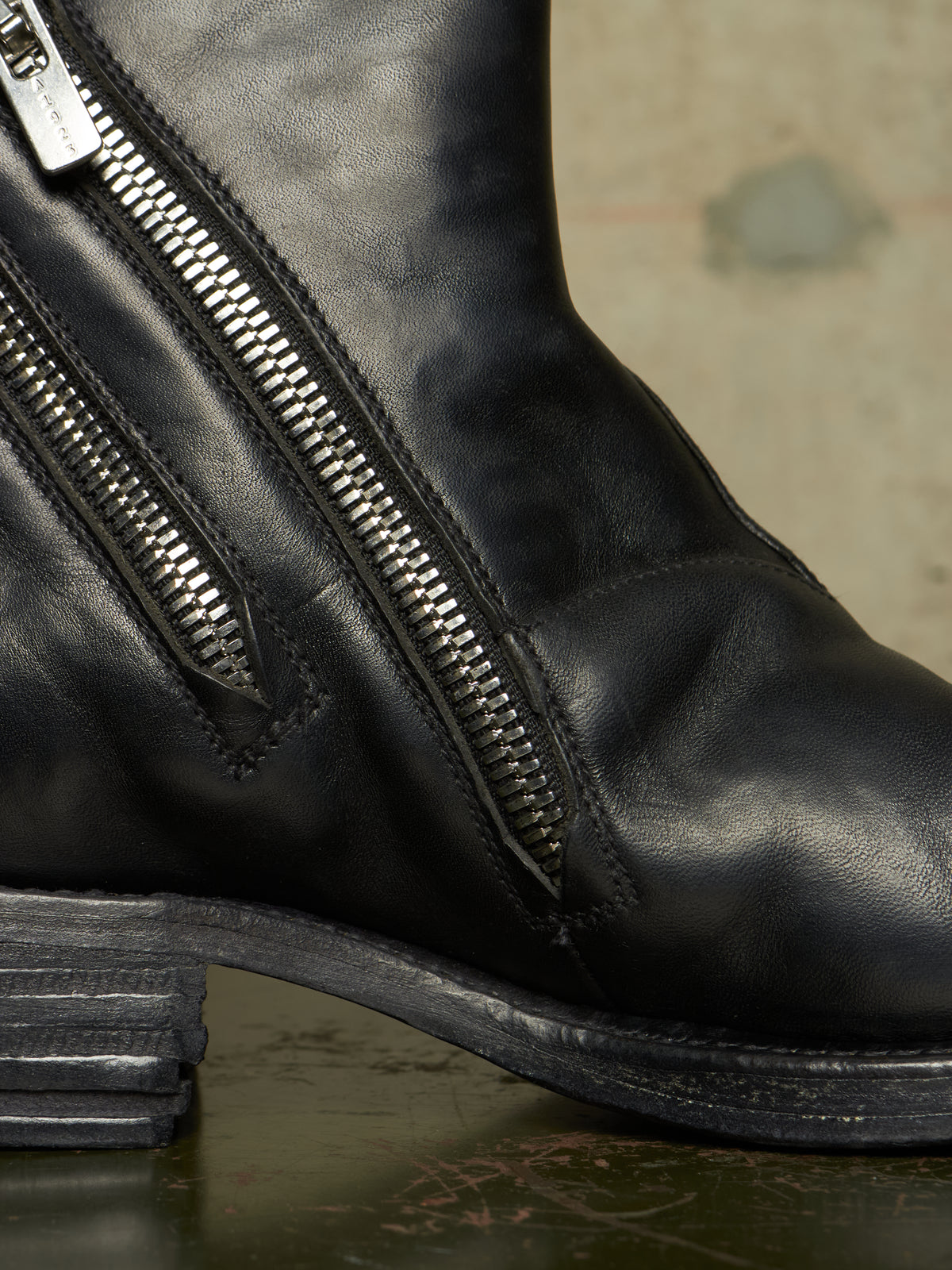 Close-up of a black leather boot with dual silver side zippers, a stacked dark heel, and a textured neutral background.