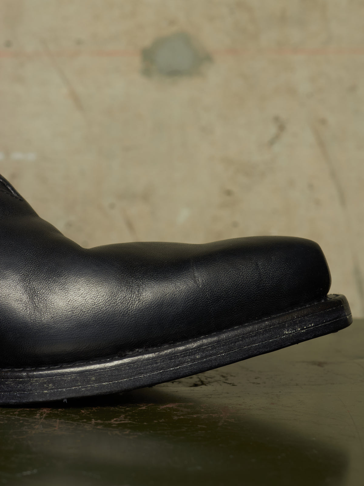 A close-up shot of a black leather boot toe showing the side profile against a textured, warm-toned concrete background.