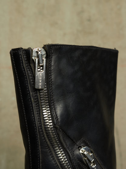 Close-up of a black leather boot featuring dual silver metal zippers, one with a decorative pull tab engraved with 'SANTO SEVERO'.