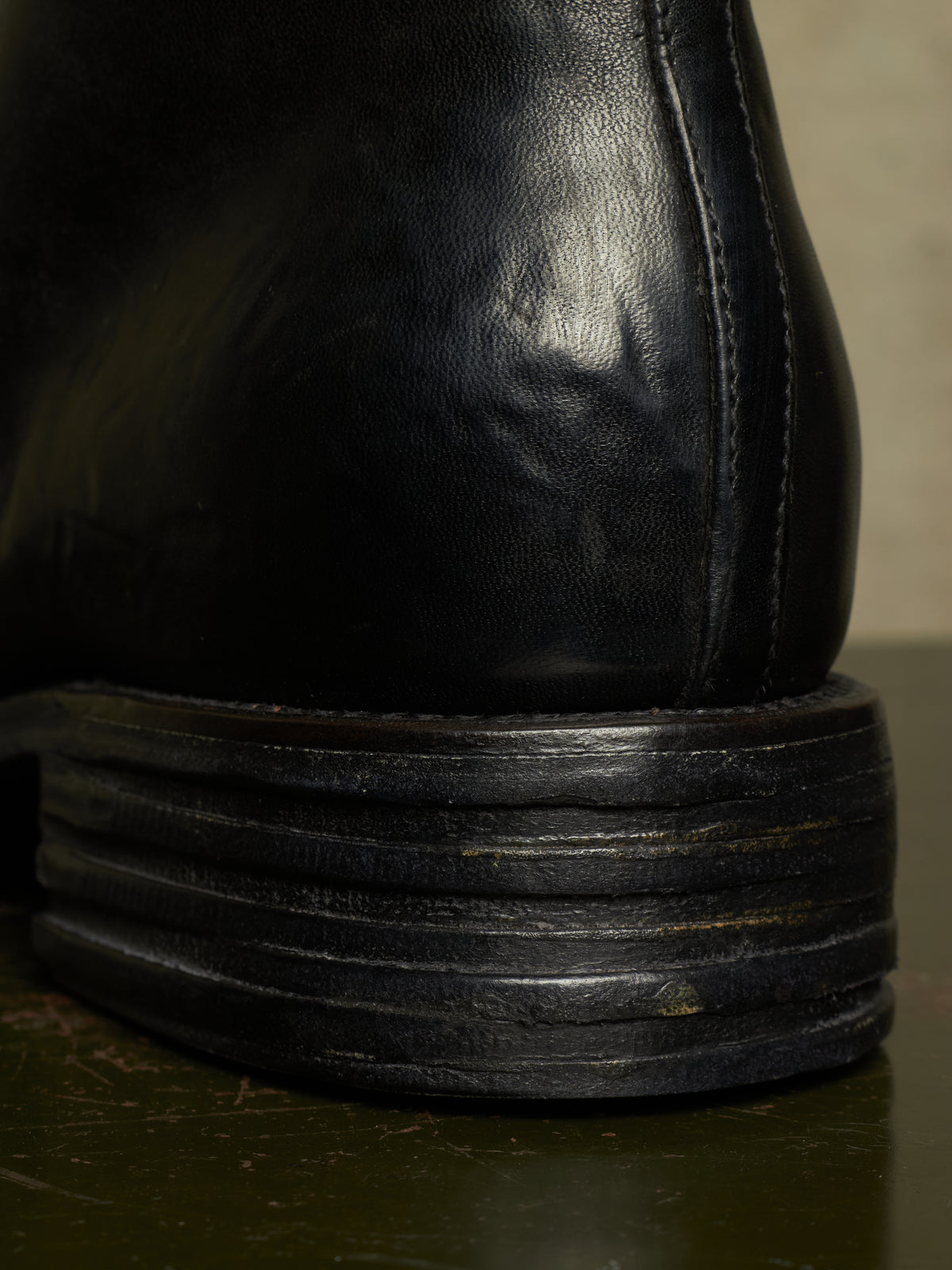 Close-up detail of the heel of a black leather boot, showing the stitched corner seam and a thick, layered black rubber sole on a dark surface.