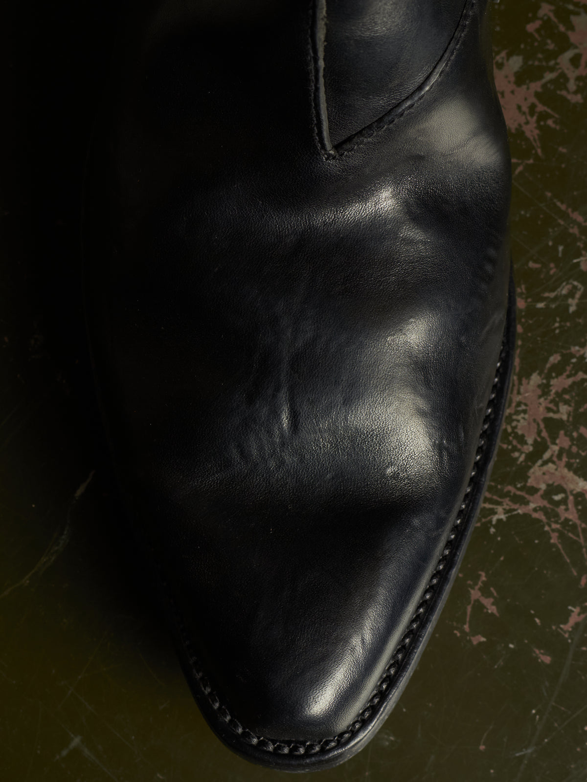 A close-up of a black leather boot toe showing the smooth texture, stitching, and a pointed tip, resting on a dark textured surface.