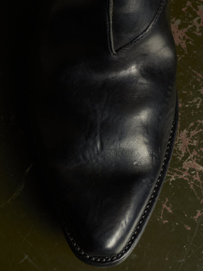 A close-up of a black leather boot toe showing the smooth texture, stitching, and a pointed tip, resting on a dark textured surface.