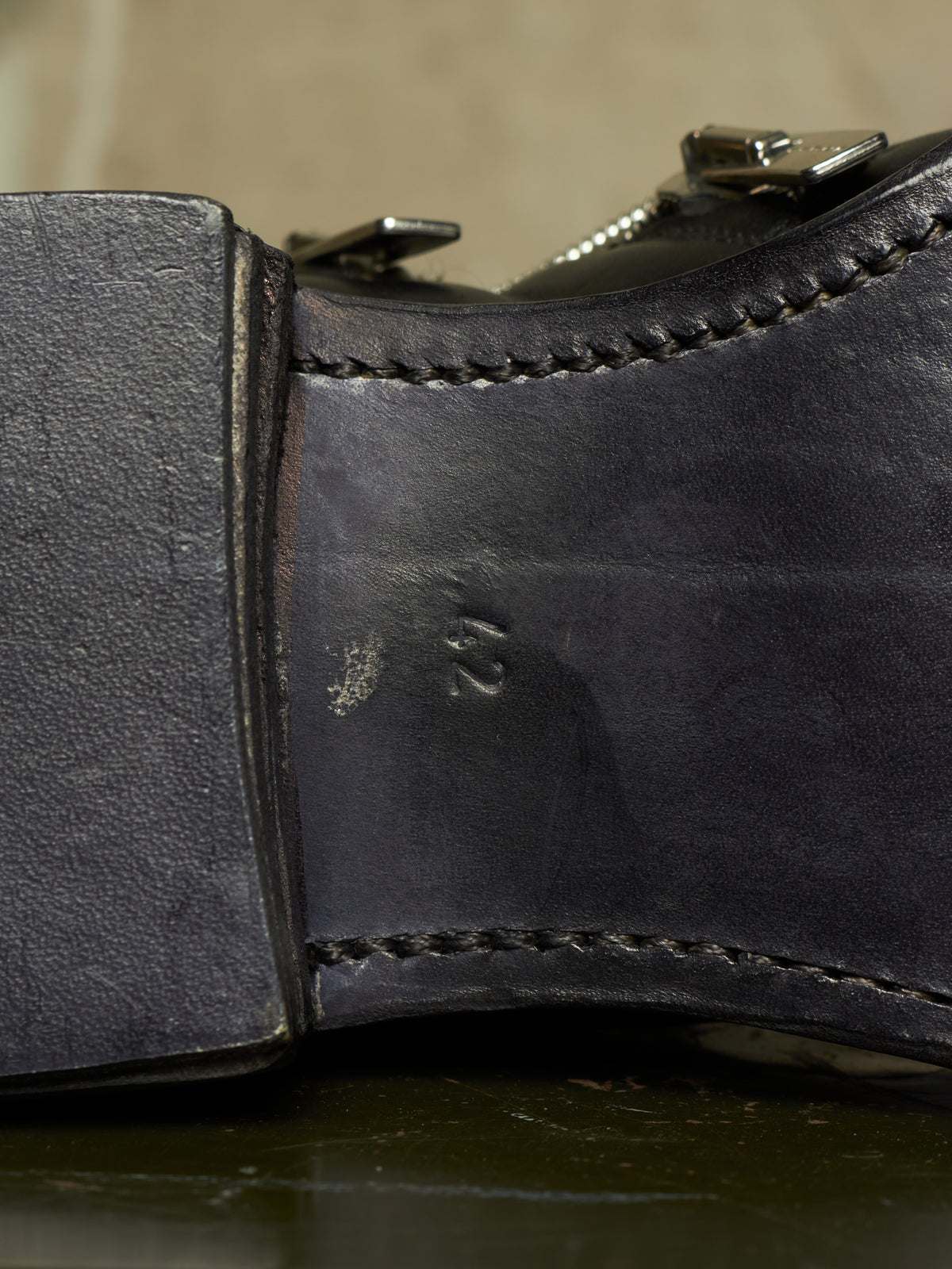 Close-up of the heel and side of a dark leather boot, showing stitched edges, a metal buckle, and an embossed size marking on the insole.