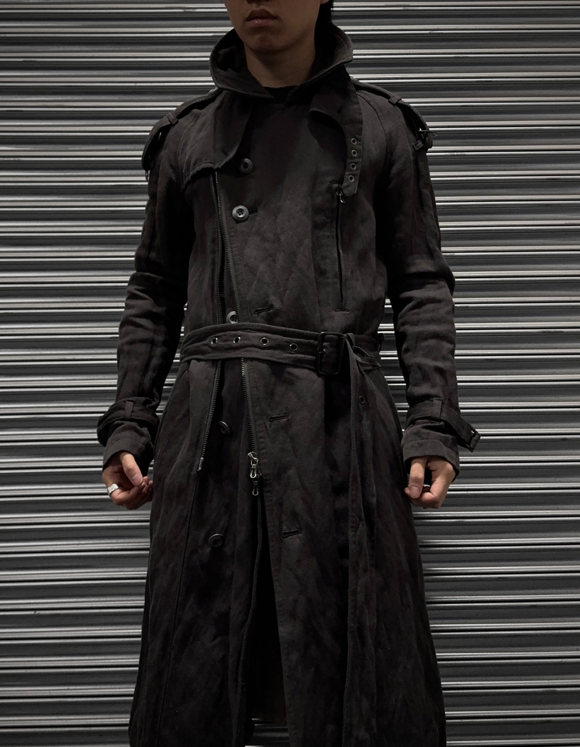 A person in an ATTACHMENT by Kazuyuki Kumagai Raglan Doctor Trench Coat stands before a closed metal shutter, their face partly shadowed and hands resting at their sides.