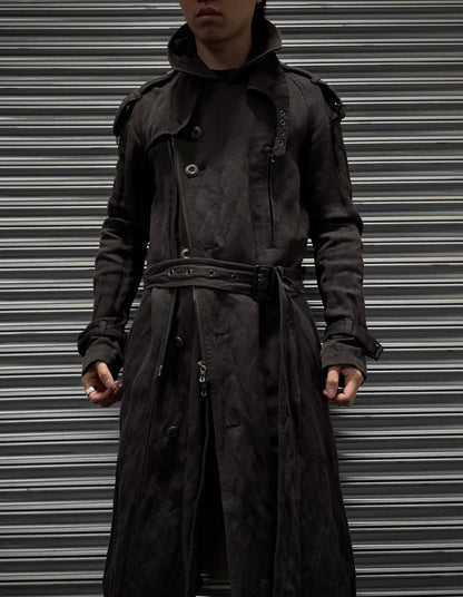 A person in an ATTACHMENT by Kazuyuki Kumagai Raglan Doctor Trench Coat stands before a closed metal shutter, their face partly shadowed and hands resting at their sides.